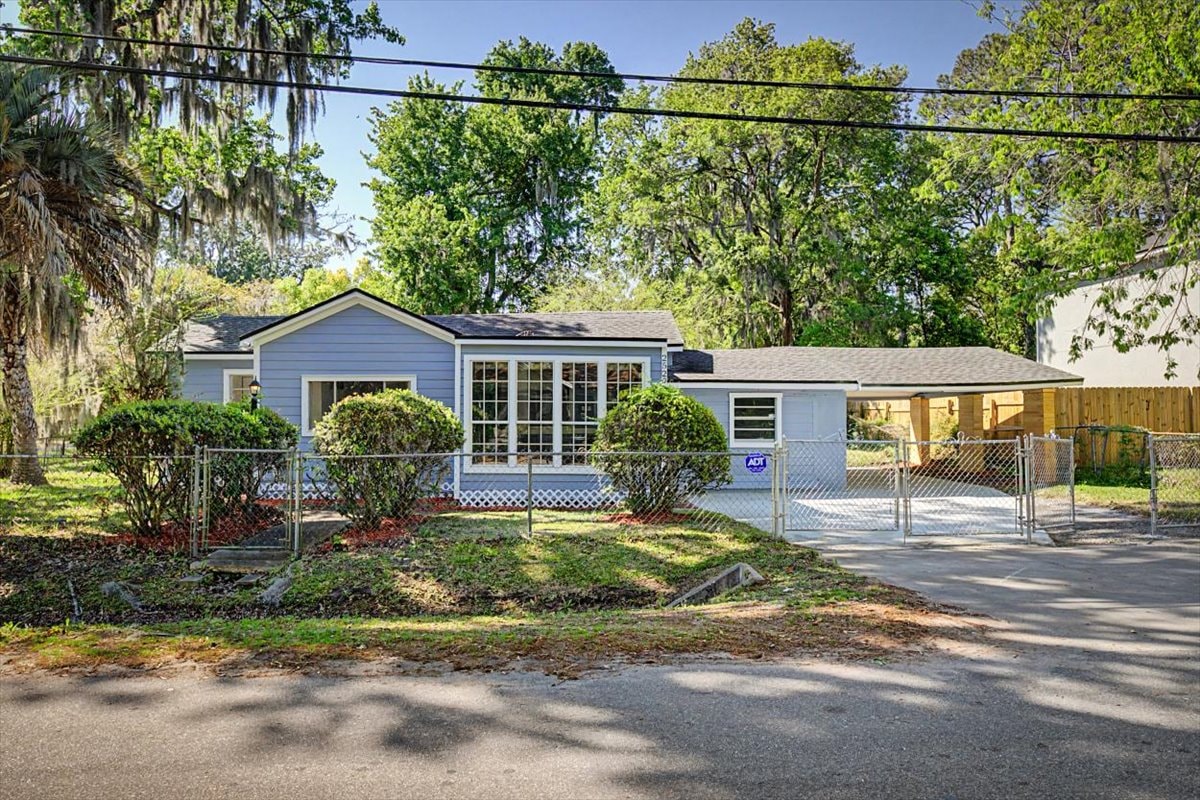 Street-level front view – blue-grey cottage with bay windows, carport, chain-link gate and mature oaks